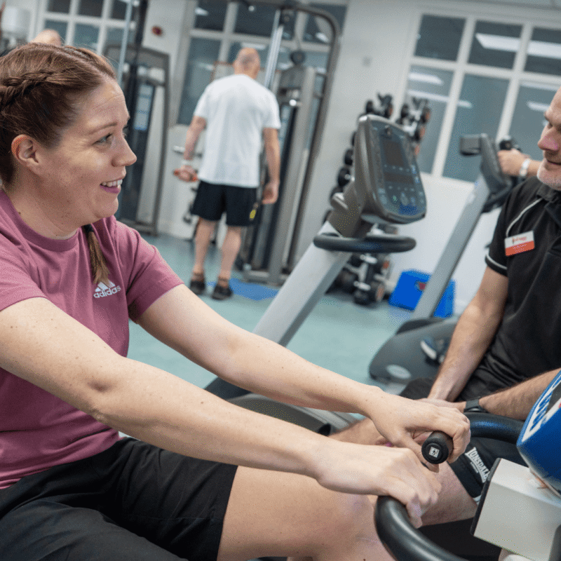 a woman using an exercise machine in a gym with a physiotherapist from the Fire Fighters charity