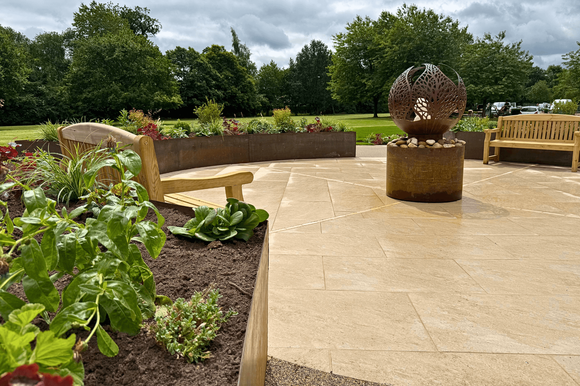 Sensory garden designed and built by Frilu for The Fire Fighters Charity at Jubilee House in Penrith. Features include curved corten steel raised beds, lush planting, natural stone paving, resin-bound gravel paths, wooden benches, and a sculptural fire pit water feature, all set against a backdrop of green lawns and trees.