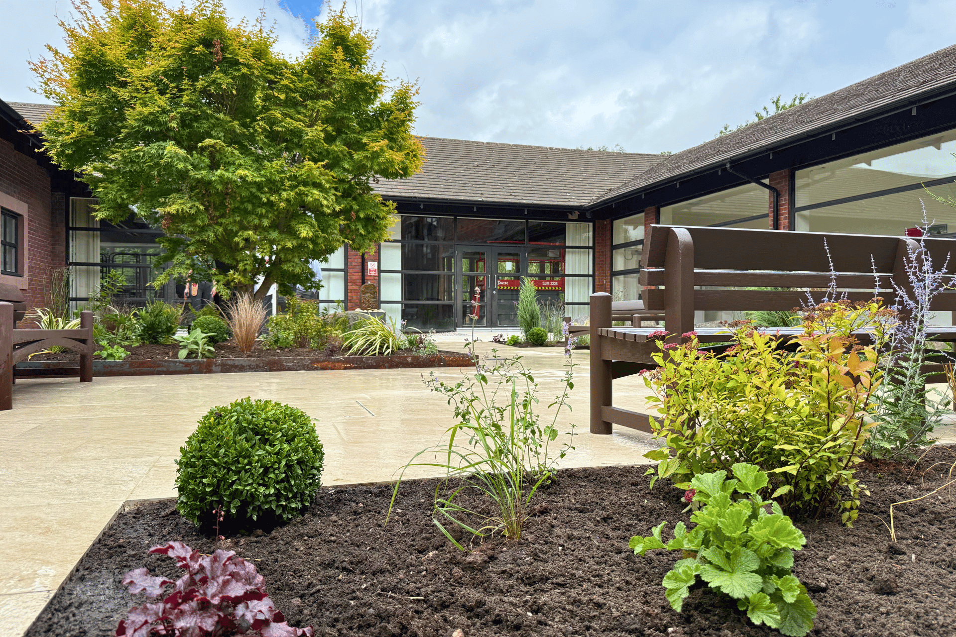 The courtyard garden at Jubilee House, Penrith for the Fire Fighters Charity. Showcasing benches, large tree amongst lush green planting and a natural stone paving walkthrough.