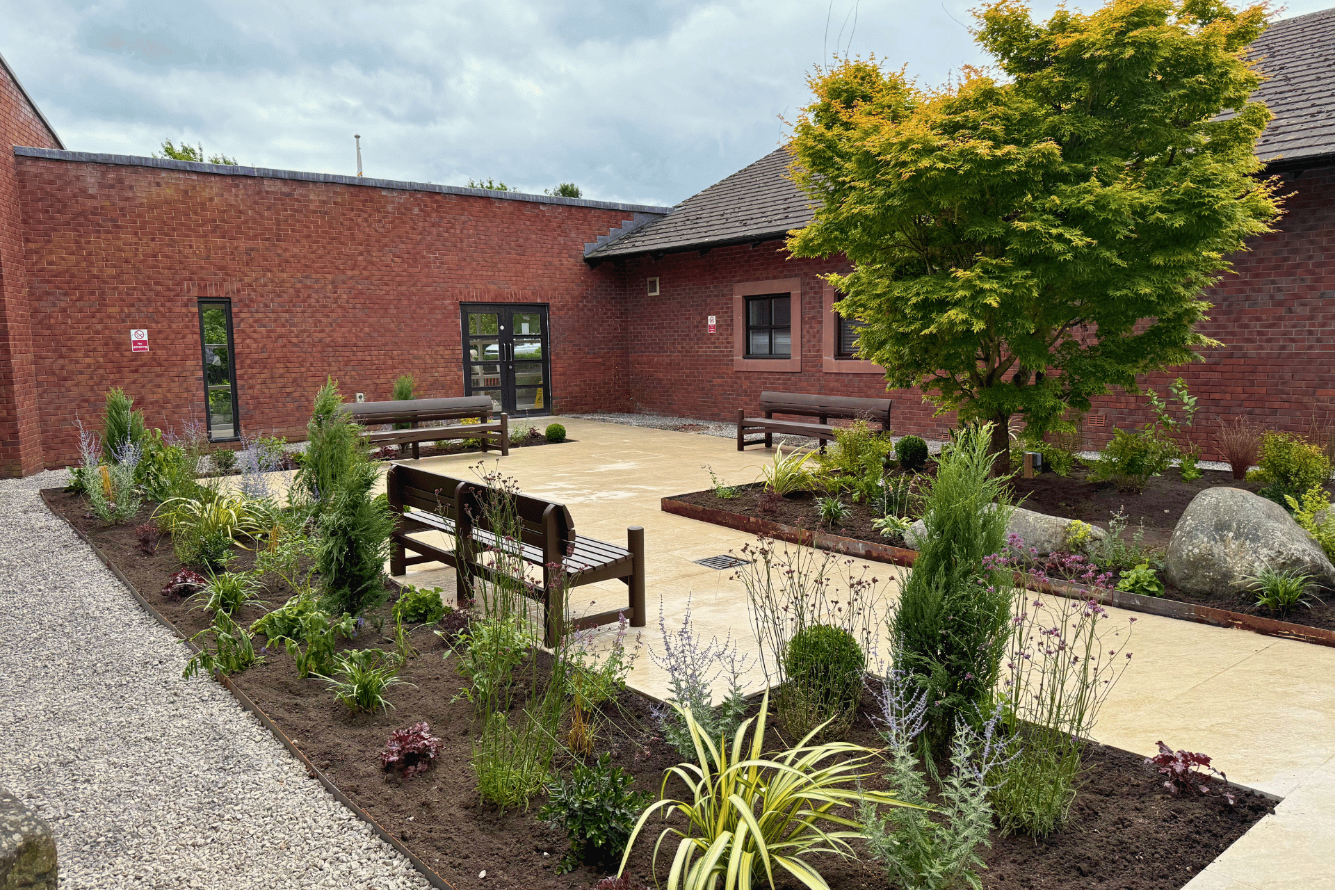 The courtyard garden at Jubilee House, Penrith for the Fire Fighters Charity. Showcasing benches, large tree amongst lush green planting and a natural stone paving walkthrough.
