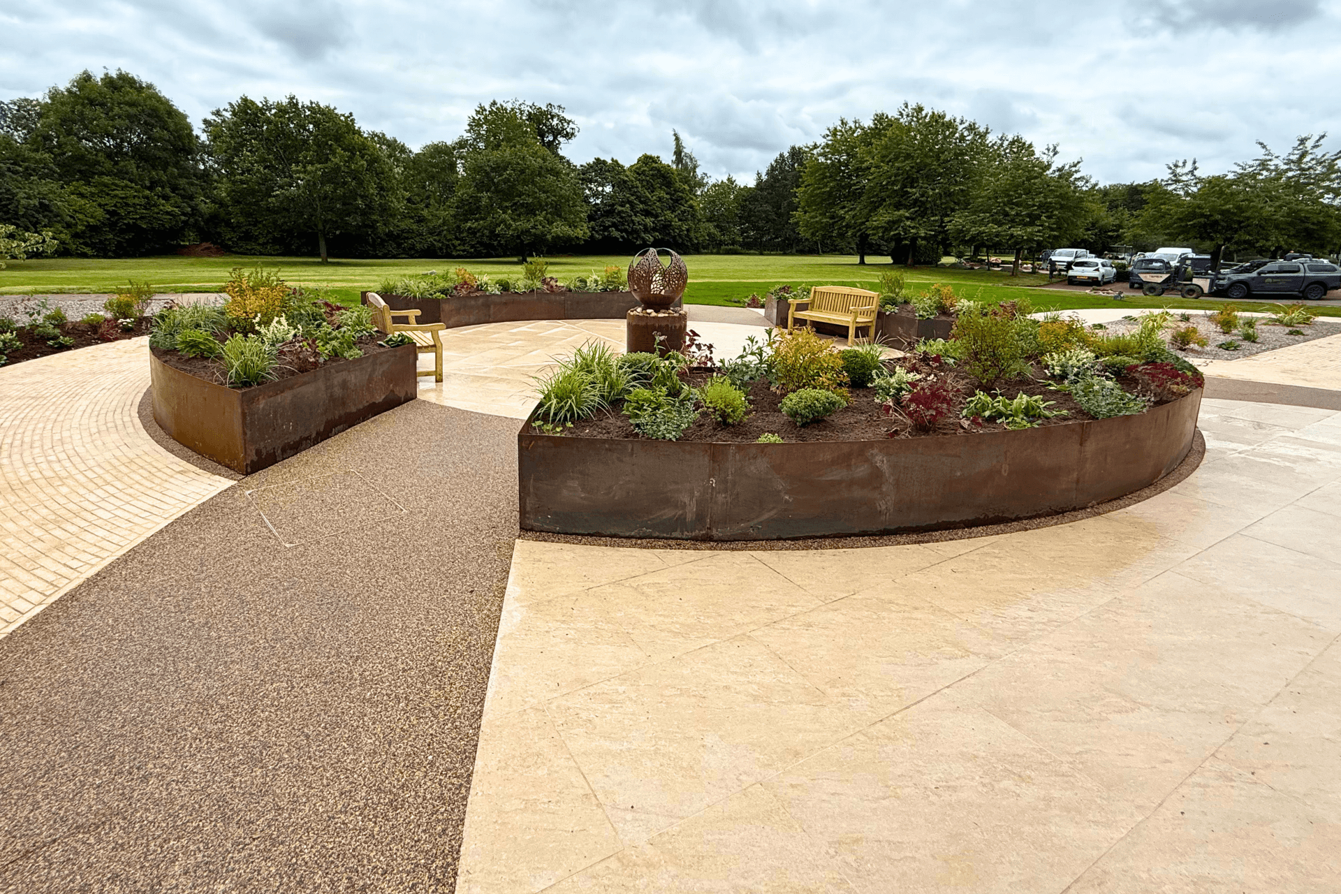Sensory garden designed and built by Frilu for The Fire Fighters Charity at Jubilee House in Penrith. Features include curved corten steel raised beds, lush planting, natural stone paving, resin-bound gravel paths, wooden benches, and a sculptural fire pit water feature, all set against a backdrop of green lawns and trees.