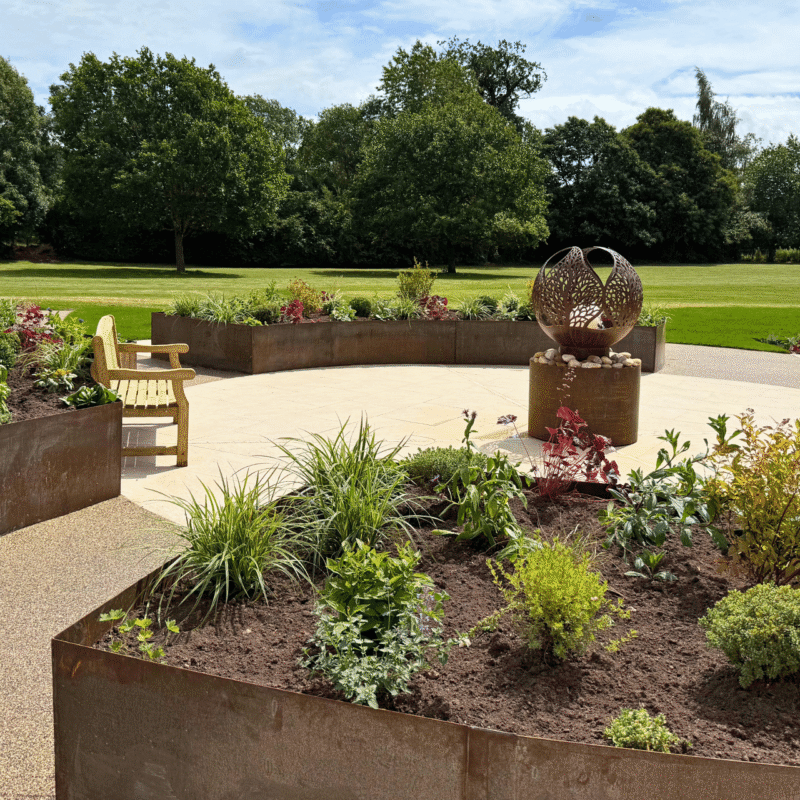 Sensory garden designed and built by Frilu for The Fire Fighters Charity at Jubilee House in Penrith. Features include curved corten steel raised beds, lush planting, natural stone paving, resin-bound gravel paths, wooden benches, and a sculptural fire pit water feature, all set against a backdrop of green lawns and trees.
