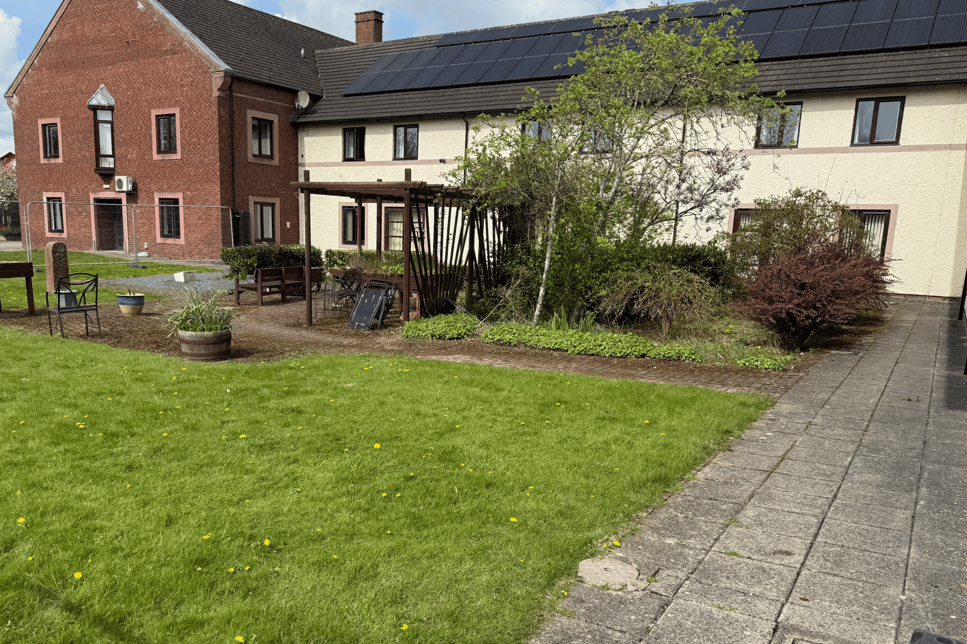 The Longest Ride garden at The Fire Fighters Charity Jubilee House in Penrith, pictured before its transformation by Frilu. The garden features a large grassy area with scattered dandelions, mature shrubs, and a worn patio space with benches, metal chairs, and a wooden pergola structure.